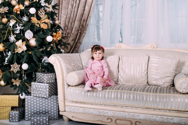 Happy girl sitting on sofa near Christmas tree indoors. Merry Christmas and Happy Holidays!