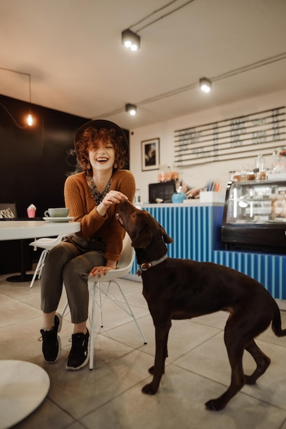 Happy girl sitting at table in cozy cafe and playing with dog looking at camera and smiling