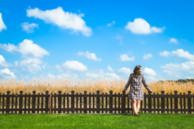 Happy girl stand with wood fence in the green field under blue cloud sky. Beautiful landscape