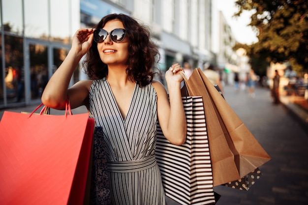 Happy girl stands near boutique shop holding a few bags with purchases.