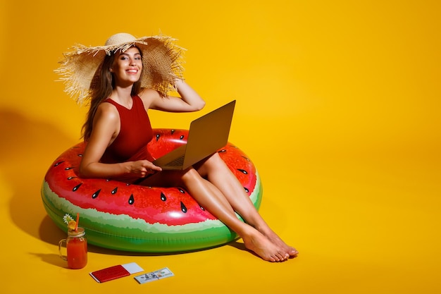 Happy girl in straw hat sits in inflatable watermelon ring uses pc on yellow background traveler