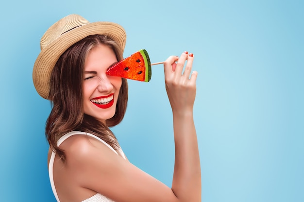 Happy girl in straw hat with a lollipop and red lipstick smiling on a pink wall . Cute girl covers her eye with lollipop fools around and laughs. Tourist girl having fun in summer