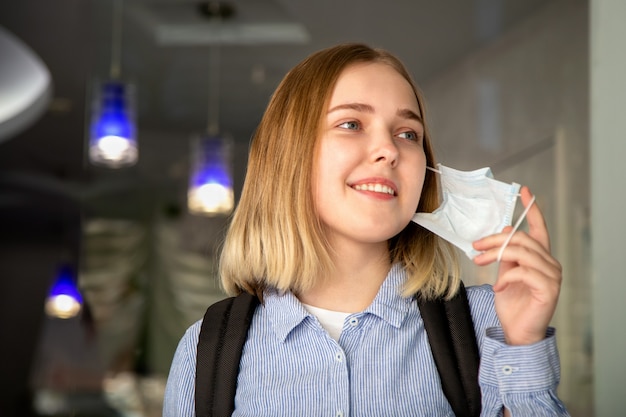 幸せな女の子の学生は、保護医療マスクを脱ぎます。コロナウイルス感染症の封鎖の終わりに大学で金髪の女子学生の肖像画。病気の後の新しい通常の安全な生活。検疫は終了しました。