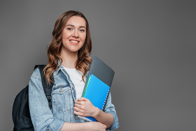 Happy girl student with backpack smiles and holds a notebook copybook folder in the hands isolated on dark grey wall . The concept of education study exams learn English