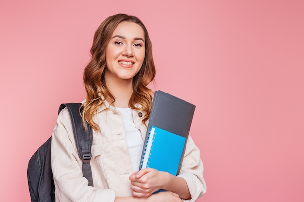 Happy girl student with backpack smiles and holds a notebook copybook folder in hands isolated on pink wall . The concept of education study exams learn English