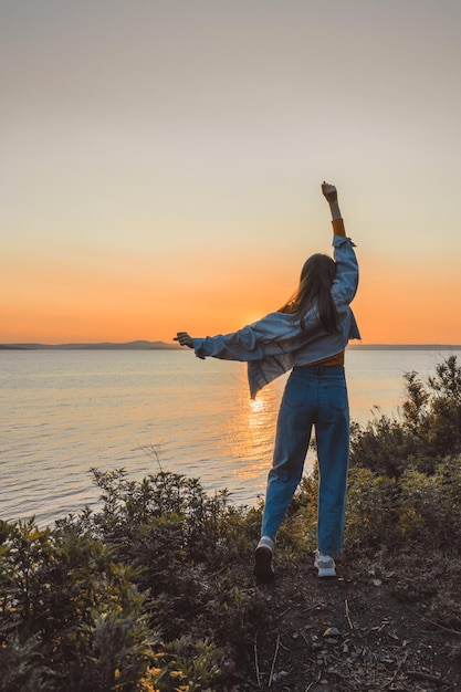 Happy girl in stylish clothes by the sea during sunset