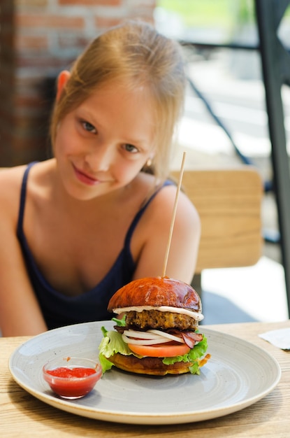 Ragazza felice, adolescente che mangia un hamburger con un tortino di carne in un ristorante in estate. fast food