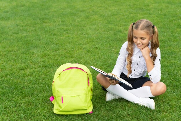 Happy girl in uniform with school bag reading book sitting on green grass