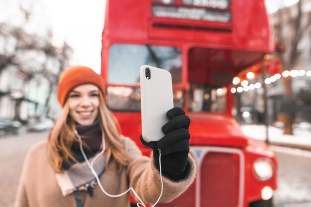 Счастливая девушка в теплой одежде стоит на улице на фоне красного автобуса и делает селфи на смартфоне