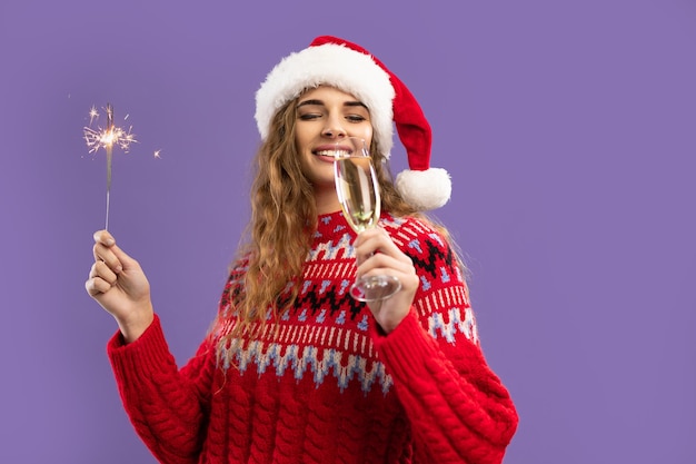 Happy girl wears santas hat drinks champagne holds sparkler on purple background christmas party