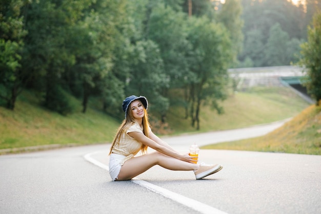 A happy girl in white shorts and a yellow blouse holds a glass of juice with a straw in her hands