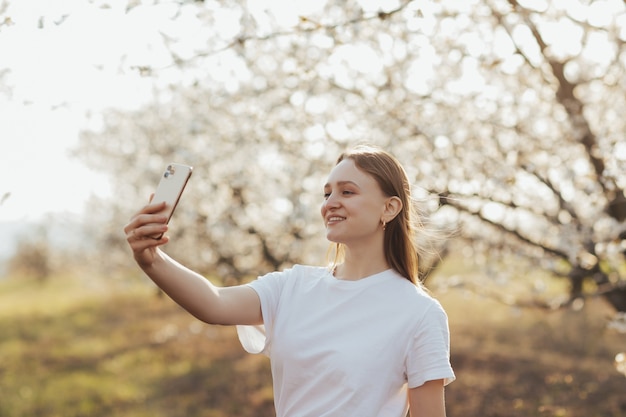 咲く木の壁に電話でselfieを取るブロンドの髪の白いTシャツの幸せな女の子。