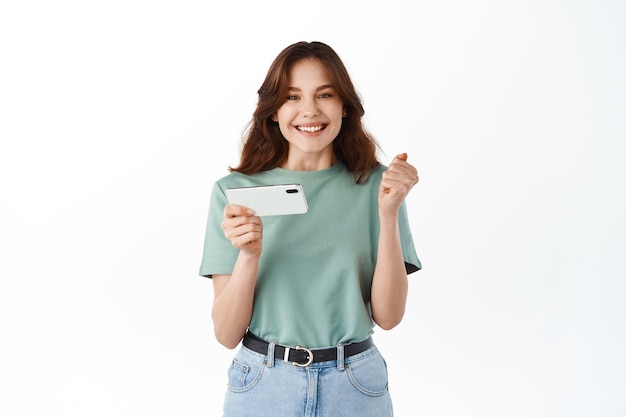 Happy girl winning on mobile video game and feeling ecstatic, rejoicing, holding smartphone horizontally, celebrating victory, standing against white wall