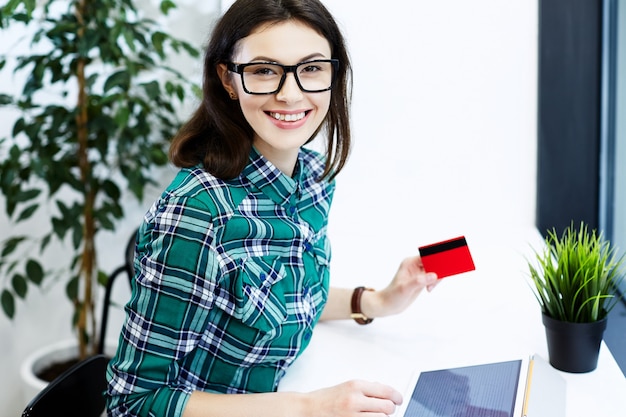 Happy girl with black hair wearing shirt and eyeglasses sitting in cafe with tablet and cup of coffee, freelance concept, holding credit card, online shopping.