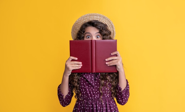 Happy girl with frizz hair hiding behind book on yellow background