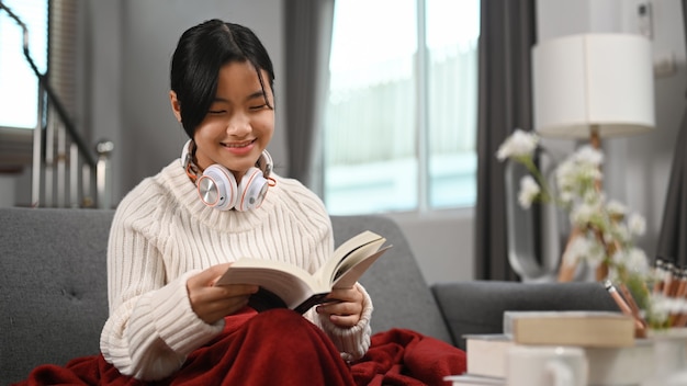 집 소파에 헤드폰으로 책을 읽고 있는 행복한 소녀.