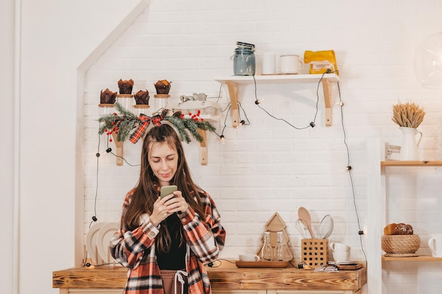 Счастливая девушка с длинными волосами на рождественской кухне смотрит в телефон