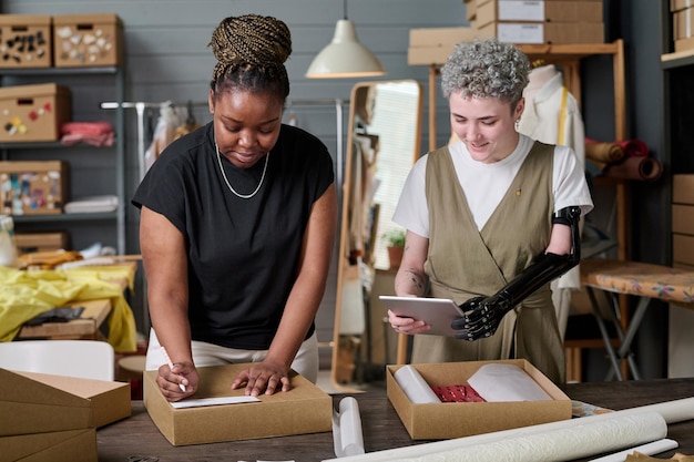 Photo happy girl with partial arm using tablet while her colleague packing parcel