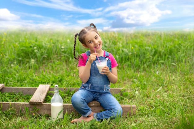 A happy girl with pigtails in denim overalls and a pink T-shirt, holds a glass of milk with a pink straw, sits on a wooden ladder, in a field on the grass. Blue sky with clouds. Copy space