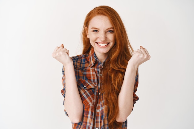 Photo happy girl with red hair feeling like winner clench hands and smiling pleased winning prize and triumphing standing satisfied against white background
