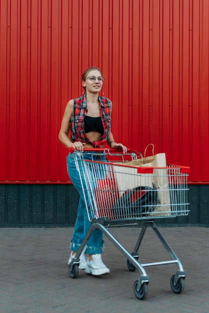 Foto ragazza felice con il carrello della spesa sul fondo rosso del negozio della parete giovane donna che spinge un carrello della spesa pieno