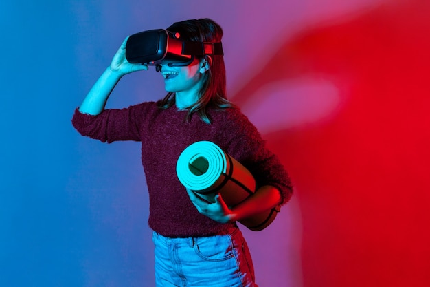 Happy girl with VR glasses headset holding rolled yoga mat, watching fitness courses