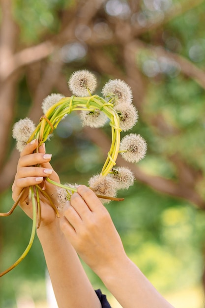 Happy girl with wreath of white dandelions on green lawn on summer sunny day