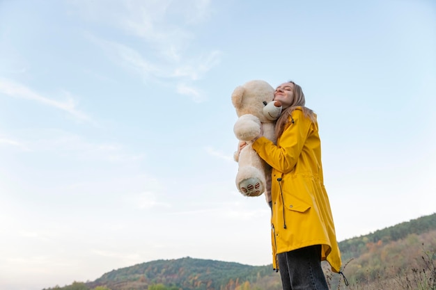 Happy girl in yellow park jacket tightly hugs soft bear toy stands on mountains background Young woman with teddy bear