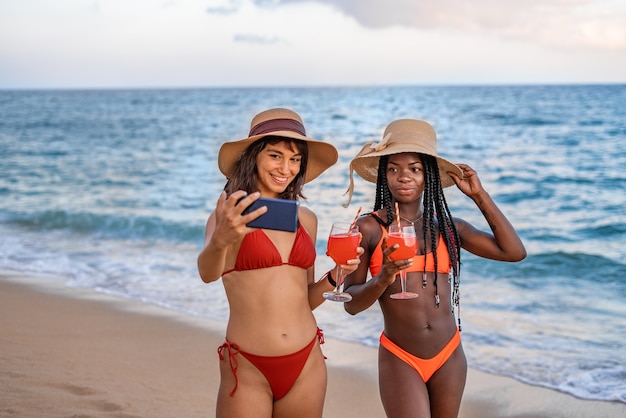 Amiche felici con cocktail che prendono selfie vicino al mare