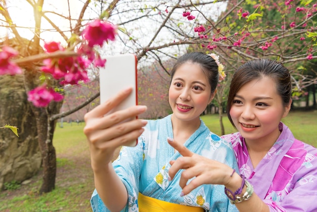 着物を着た幸せなガールフレンドの女性は、モバイルスマートフォンのセルフィーで写真を撮り、日本の桜の庭を旅して咲く桜の花を指差すことに焦点を当てています。