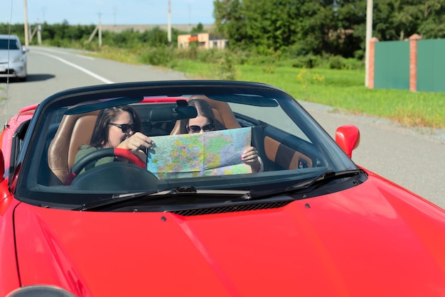 Happy girls in sunglasses driving red cabriolet car during vacation road trip having fun together discovering new places looking to the map Road trip travel enjoying freedom concept Selective focus