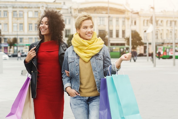 Happy girls with shopping bags outdoors. Smiling female friends in bright warm casual clothes having a city walk. Friendship, urban lifestyle and leisure concept