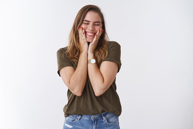 Happy good-looking positive sincere young european woman chestnut hair olive t-shirt squeez cheeks joyfilly close eyes smiling broadly white teeth grin, standing amused excited white background