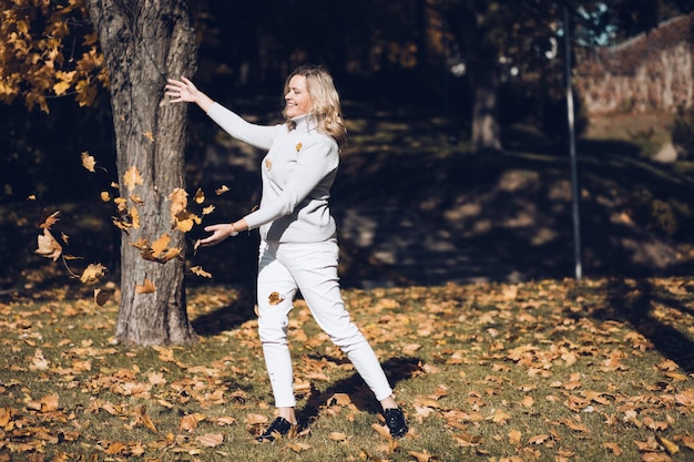 Happy good-looking young fair-haired woman dressed in beige jacket and white jeans playfully throwing leaves outdoors in park or forest in warm autumn days possibly in Septemper or October