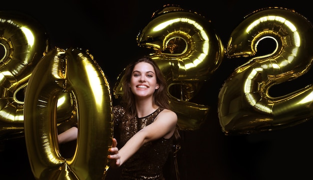 Счастливая великолепная девушка в стильном сексуальном праздничном платье с золотыми воздушными шарами 2022 года, веселится на вечеринке в канун Рождества или Нового года.