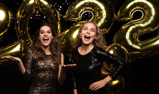 Happy gorgeous girls in stylish sexy party dresses holding gold 2022 balloons, having fun at Christmas or New Year's Eve Party.