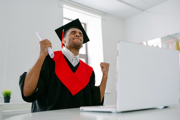 Felice di laurearsi, uno studente nero si siede a un laptop con un abito e un berretto quadrato e comunica con i compagni laureati cerimonia virtuale di laurea e convocazione grag nello spazio della copia della stanza luminosa