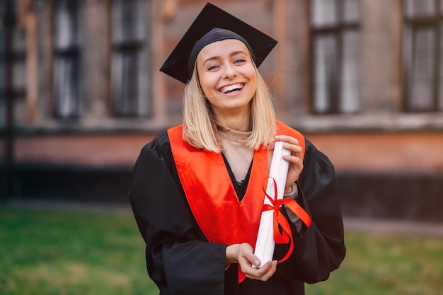 A happy graduate stands in a university garden in a mantle with a diploma in her hand smiles and looks at the camera