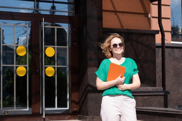 A happy graduate student girl in sunglasses holding a digital tablet book noutbook against the university building