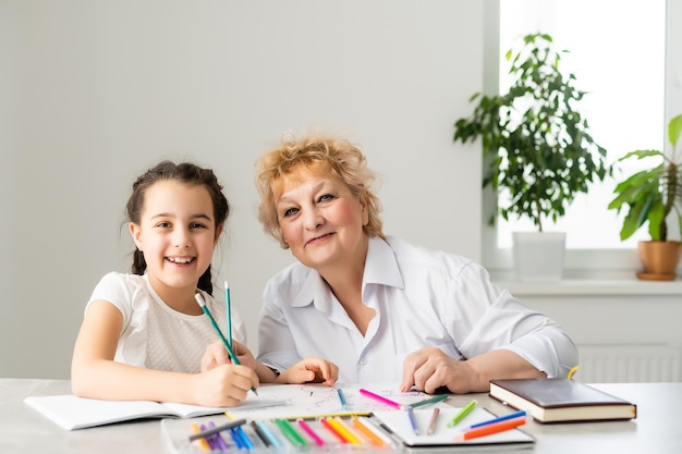 Happy grandchild with grandmother having fun, drawing colored pencils, sitting together at home, laughing preschool girl with smiling grandma painting picture, playing with granddaughter.