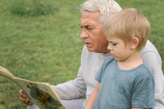 Happy grandfather reading book to curious grandson outdoors Close up Grandpa with grandchild spending time together Family time comes in various forms 4s year boy and senior man read story in park