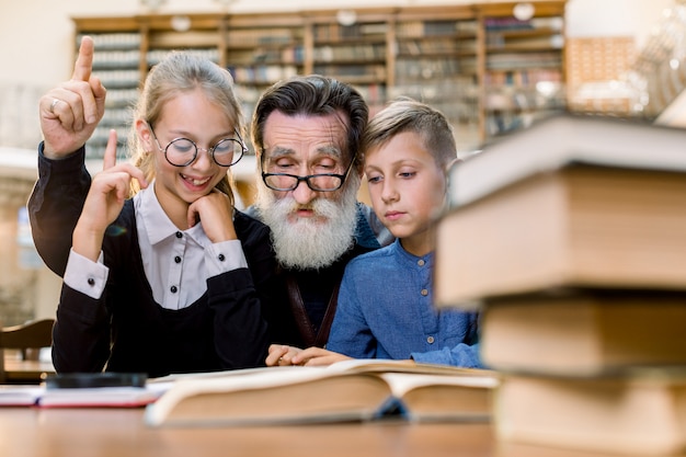 Happy grandfather reading book with grandson and granddaughter, sitting at the table in old vintage library. elderly man and the girl are pointing their fingers up and laughing