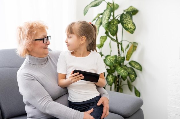 Happy grandmother and granddaughter using phone together, sitting on cozy sofa at home, browsing mobile device apps, grandma with grandchild playing game, looking at screen, having fun.