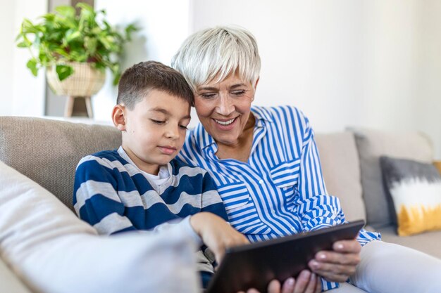 Happy grandmother and grandson using tablet together, sitting on cozy sofa at home, browsing tablet device apps, grandma with grandchild playing game, looking at screen, having fun