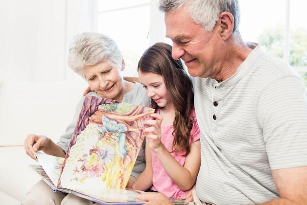 Счастливые бабушка и дедушка с внучкой читают книгу на диване