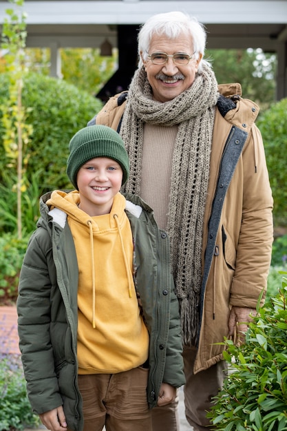 Happy grandson and grandfather in warm jackets and other casuawlear standing by each other in front of camera against green bushes