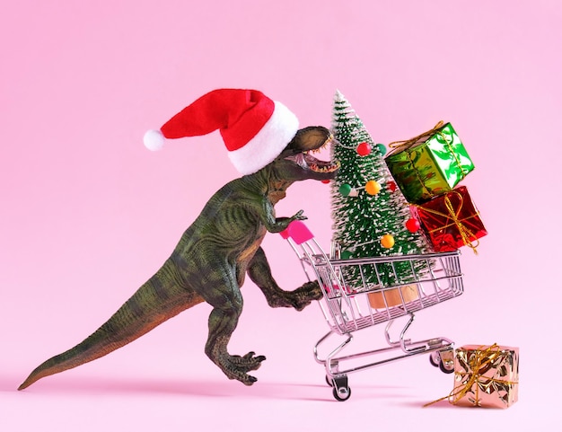 Happy green dinosaur in Santa Claus hat carrying shopping cart with Christmas tree and gift boxes on pastel pink background