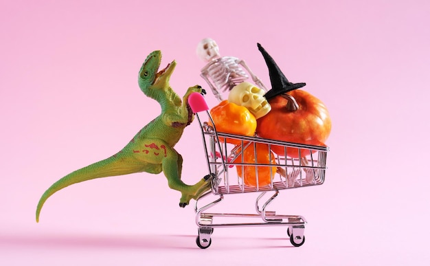 Photo happy green dinosaur with shopping trolley full of halloween decorations on pink background