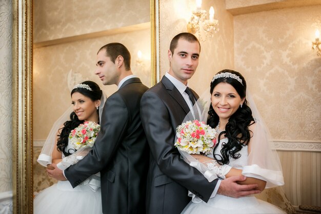 Happy groom and the bride stand near a mirror and are reflected in it