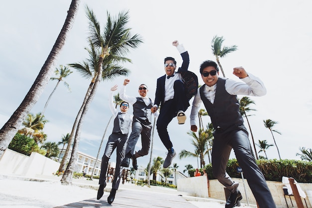 Happy groom and groomsmen jump on the way along the palms